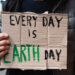 closeup of a young caucasian man, on the street, showing a brown cardboard signboard with the text every day is Earth day handwritten in it