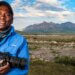 african american man with camera in his hand smiling at the camera. Mountain and a beautiful field in the background.