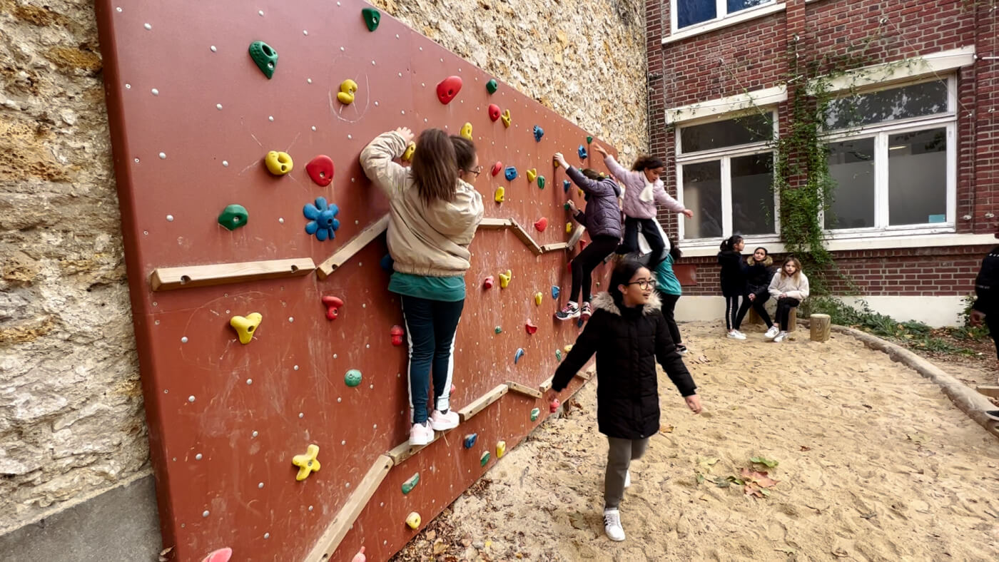 Four girls climb on a short climbing wall fixed to the side of a school.