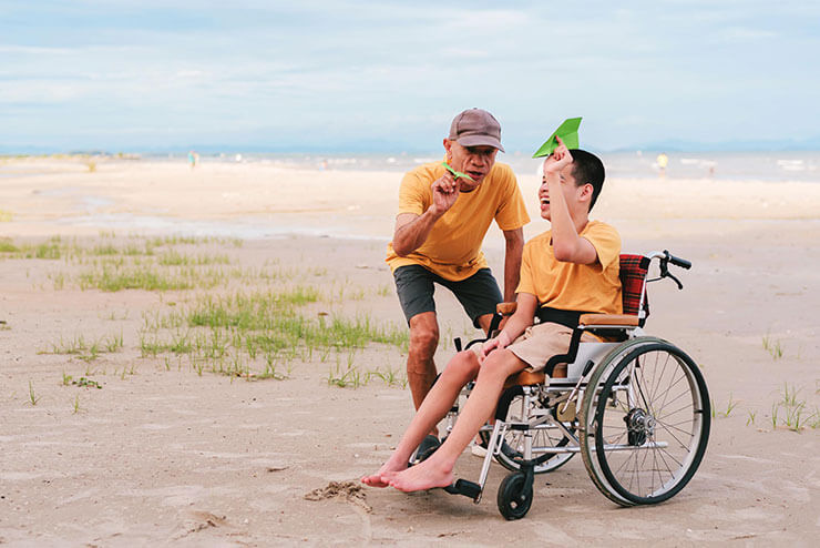 An elderly man and a young boy with special needs are enjoying a day at the beach. The man, wearing a cap and a yellow shirt, is crouched down beside the boy, who is sitting in a wheelchair. The boy, also dressed in a yellow shirt, is holding a green paper airplane and looking at it intently. They are on a sandy beach with patches of green grass, and the ocean is visible in the background. The sky is clear with a few clouds, creating a peaceful and bright atmosphere.