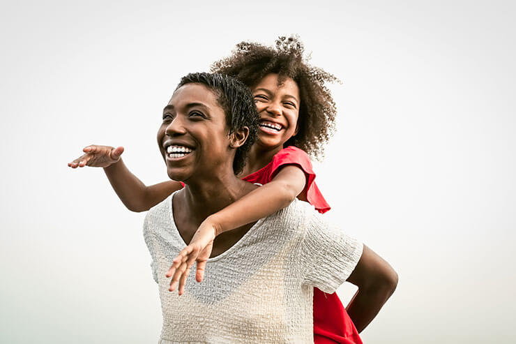 A joyful moment between a black mother and her daughter. The mother, dressed in a white top, is giving her young daughter a piggyback ride. Both are smiling broadly and looking into the distance. The daughter, wearing a red dress, has her arms outstretched and her curly hair bouncing in the air.
