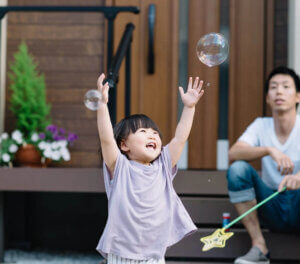 child playing with bubbles and father sitting nearby watching